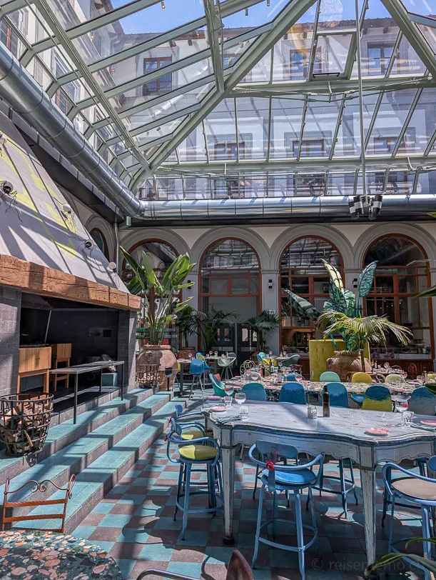 Helles 25hours Hotelrestaurant mit Glasdach, türkisfarbenem Fliesenboden, bunten Stühlen und tropischen Pflanzen in einem historischen Gebäude mit Rundbogenarkaden in Florenz