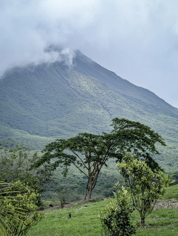 Vulkankegel des Arenal ragt aus üppiger grüner Tropenvegetation empor, während Wolken um seinen Gipfel ziehen