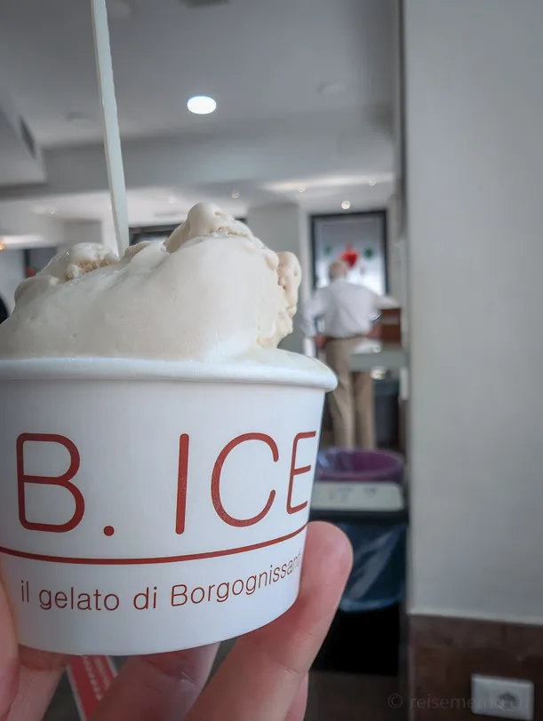 Eine Hand hält einen weissen Gelato-Becher mit der Aufschrift B.ICE – il gelato di Borgognissanti, gefüllt mit cremigem hellem Glacé und einem Löffelstiel, im Hintergrund das Innere der Gelateria