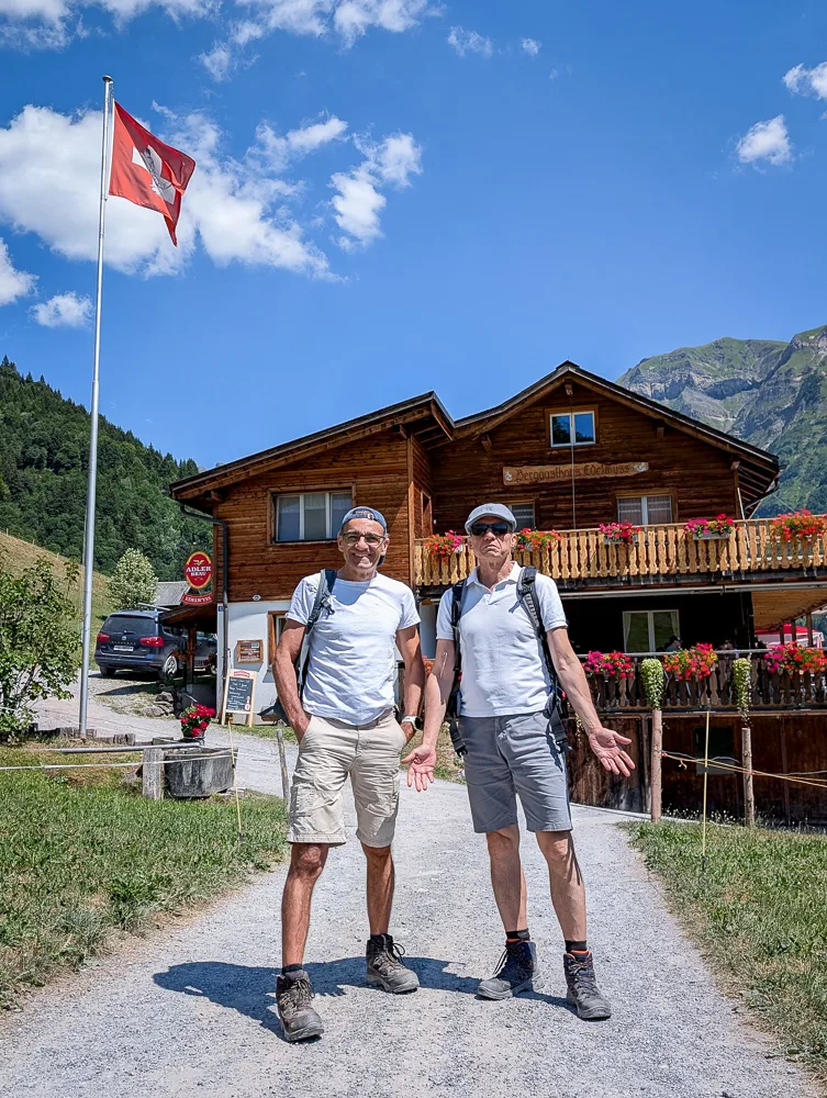 Zwei Wanderer stehen auf einem Kiesweg vor einem Holzchalet mit Schweizer Flagge in alpiner Landschaft