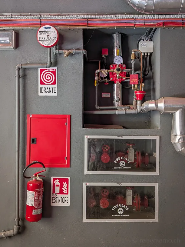 Graue Wand mit rotem Feuerlöscher, Idrante-Schild, Fire-Glass-Schaukästen, Sprinkler-Alarm-Schild und rotem Leitungssystem im 25hours Hotel in Florenz