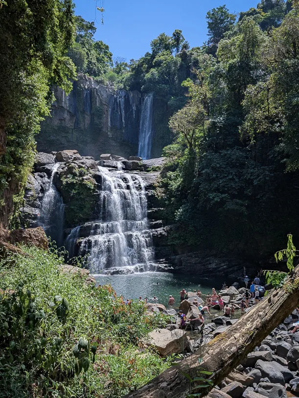 Mehrstufiger Wasserfall mit natürlichem Pool am Fuss, umgeben von dichtem tropischem Regenwald und Besuchern auf den Felsen
