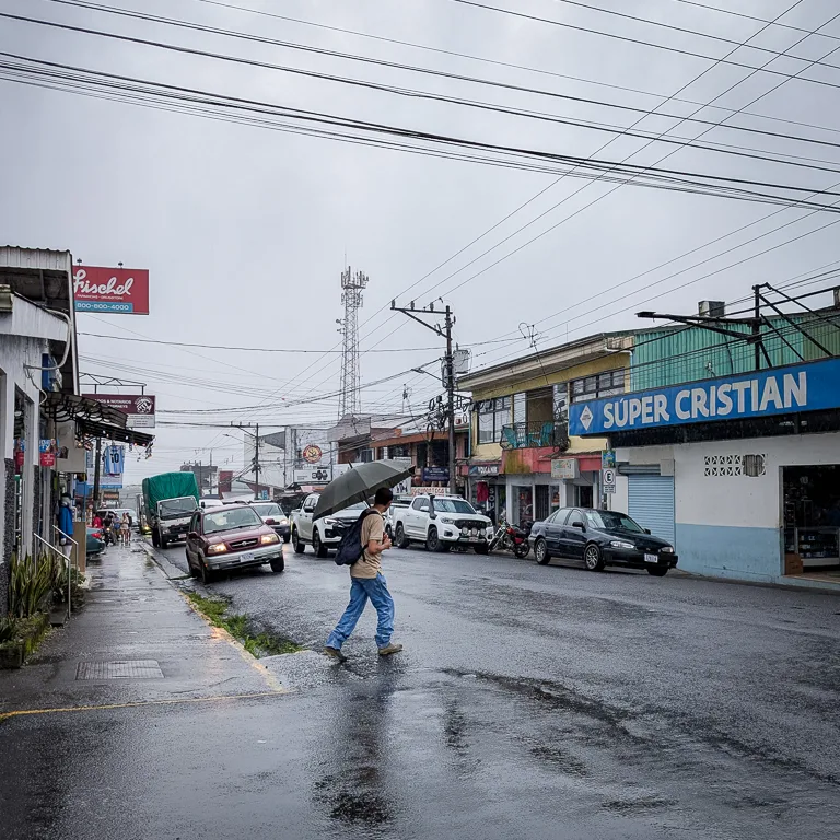 Nasse Strasse in La Fortuna, einer costa-ricanischen Kleinstadt mit Geschäften, parkierten Autos und einer Person die bei Regen über die Strasse geht