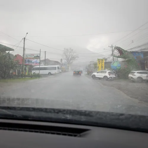 Blick durch eine regennasse Windschutzscheibe auf eine Dorfstrasse in Costa Rica mit parkierten Fahrzeugen und Geschäften bei starkem Regen
