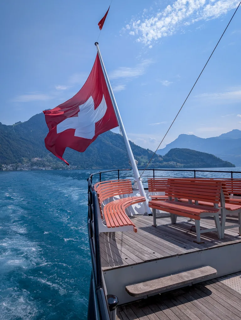 Heck eines Ausflugsschiffes mit Schweizer Fahne und leeren Bänken auf dem Vierwaldstättersee, umgeben von Bergen