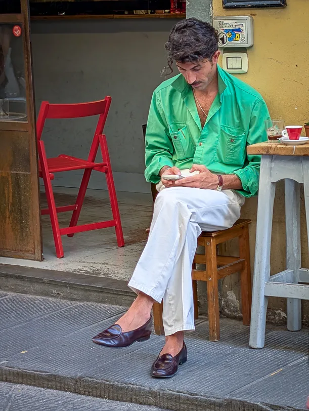 Ein elegant gekleideter Mann im grünen Hemd und weissen Hosen sitzt auf einem Hocker vor einem Café in Florenz und schaut auf sein Smartphone