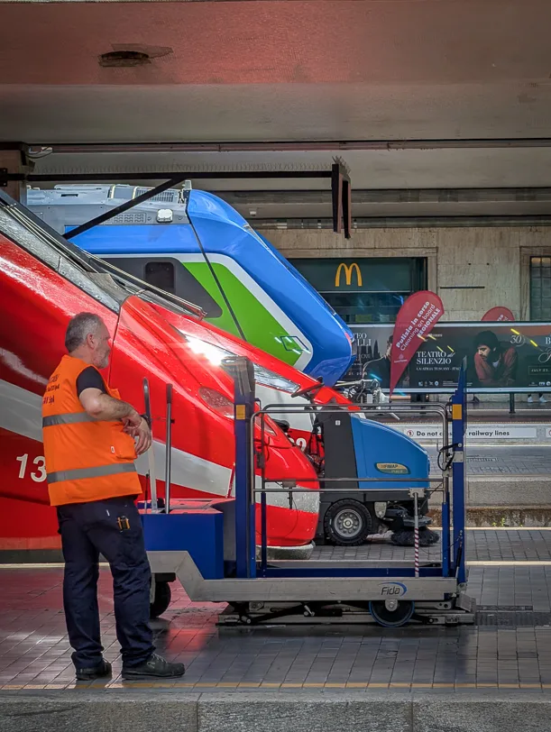 Ein Bahnmitarbeiter in orangefarbener Warnweste steht auf dem Bahnsteig vor zwei roten und einem grün-blauen Trenitalia-Zug am Bahnhof Santa Maria Novella Florenz