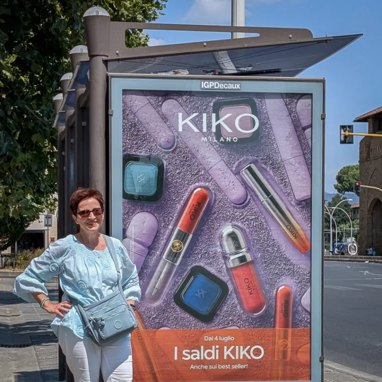 Eine Frau in hellblauer Bluse und weisser Hose lehnt an einem Werbeplakat der Kosmetikmarke Kiko Milano an einer Bushaltestelle in Florenz