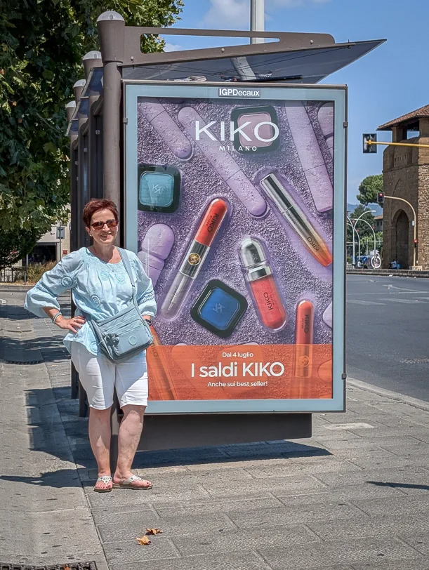 Eine Frau in hellblauer Bluse und weisser Hose lehnt an einem Werbeplakat der Kosmetikmarke Kiko Milano an einer Bushaltestelle in Florenz