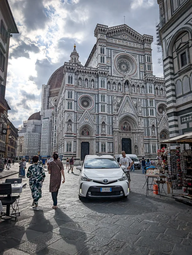 Blick auf die weiss-grün-rot marmorierte Westfassade des Florentiner Doms Santa Maria del Fiore mit der Brunelleschi-Kuppel im Hintergrund, davor Fussgänger und ein weisses Taxi auf dem Kopfsteinpflaster
