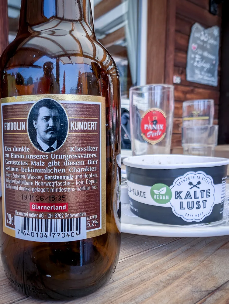 Bierflasche mit Fridolin Kundert Etikett sowie Becher mit veganer Glace auf Holztisch in Bergrestaurant