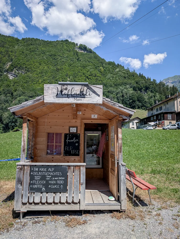 Kleines Holz-Hoflädeli mit Verkaufsbrett vor grüner Bergkulisse in Glarus Süd