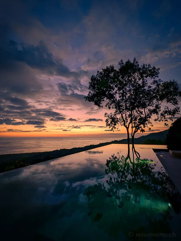 Kura Boutique Hotel Infinity Pool mit perfekter Spiegelung eines Baumes, dahinter der Pazifik und ein dramatischer Sonnenuntergangshimmel in orange und blauen Tönen