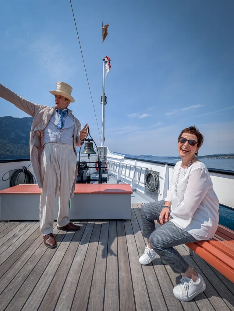 Frau lacht auf Schiffbank, daneben zeigt Mann in historischer Kleidung auf Deck des Dampfschiffs