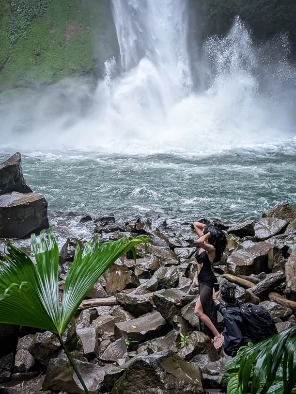 Eine Wanderin steht auf Felsblöcken vor einem hohen Wasserfall, umgeben von tropischer Vegetation und dichtem Sprühnebel