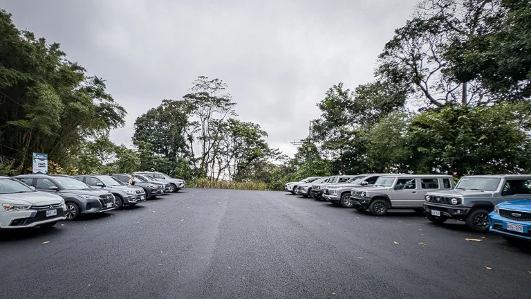 Asphaltierter Parkplatz mit vielen geparkten Autos umgeben von dichtem tropischen Regenwald unter bewölktem Himmel