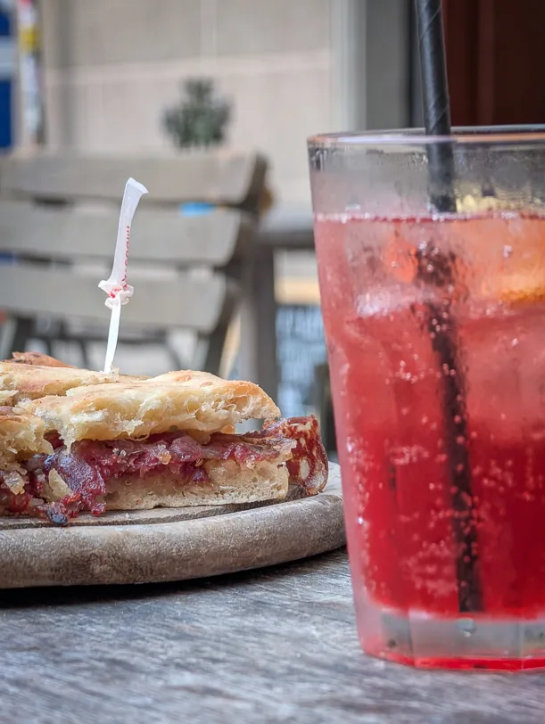 Ein aufgeschnittenes Panino mit dunkler Fleischfüllung auf einem Holzteller neben einem roten Sprudel-Getränk mit Strohhalm auf einem Terrassentisch des 25hours Hotels in Florenz