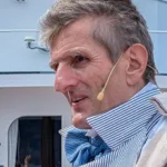 Ein Schauspieler in historischer Kleidung mit Mikrofon steht auf dem Deck eines Dampfschiffs, im Hintergrund eine uniformierte Frau
