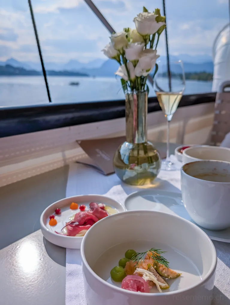 Fein angerichtetes Gourmetgericht und Kaffee auf einem weissen Tisch mit Seeblick, im Hintergrund Blumen in Vase und Sektglas