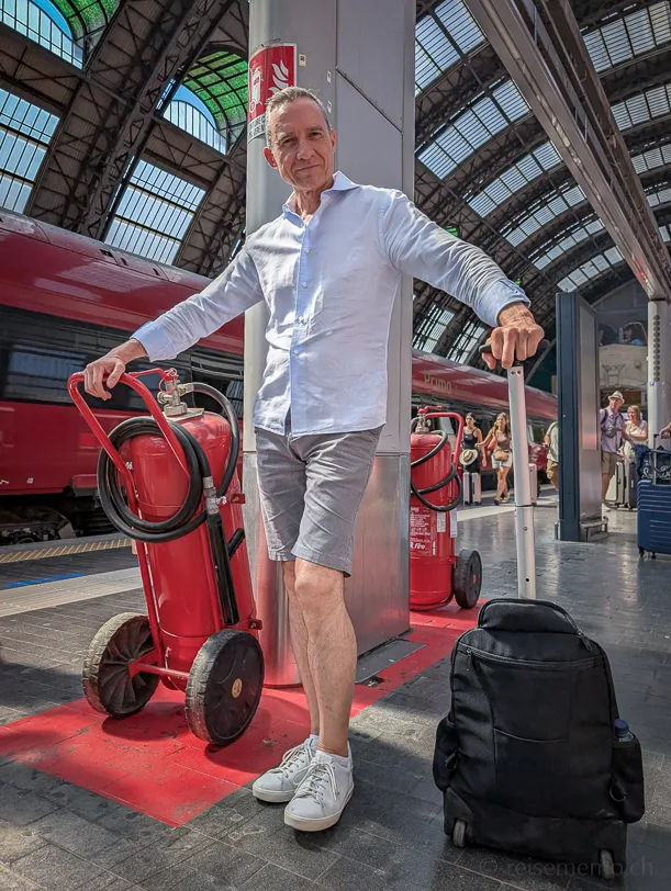Walter Schärer in weissem Hemd und grauen Shorts lehnt lässig an einer Säule auf dem Bahnsteig der Stazione Centrale in Mailand, neben ihm rote Feuerlöscher und ein schwarzes Gepäckstück, im Hintergrund ein roter Zug von Trenitalia