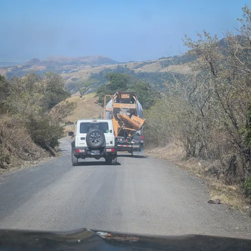 Blick auf eine Bergstrasse mit einem weissen Geländewagen und einem Lastwagen, umgeben von bewaldeten Hügeln unter blauem Himmel