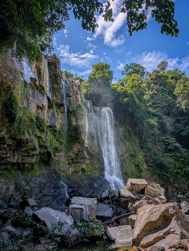 Oberer Nauyaca Wasserfall stuerzt ueber eine bewachsene Felswand zwischen tropischer Vegetation hinab auf grosse Steinbloecke.