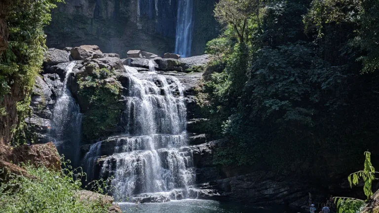 Mehrstufiger Wasserfall stürzt über Felsformationen in ein türkisblaues Becken, umgeben von dichtem tropischem Regenwald mit Besuchern am Ufer