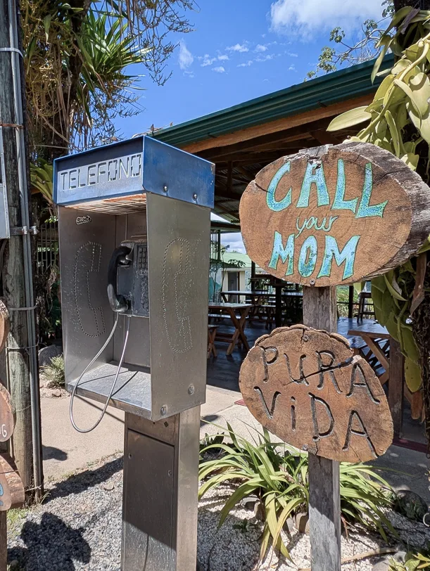 Alte Telefonzelle mit Holzschildern 'Call your Mom' und 'Pura Vida' vor tropischer Vegetation