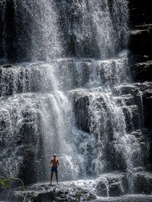 Person steht vor dem mehrstufigen Nauyaca Wasserfall mit verschiedenen Kaskaden und natuerlichen Felspools.