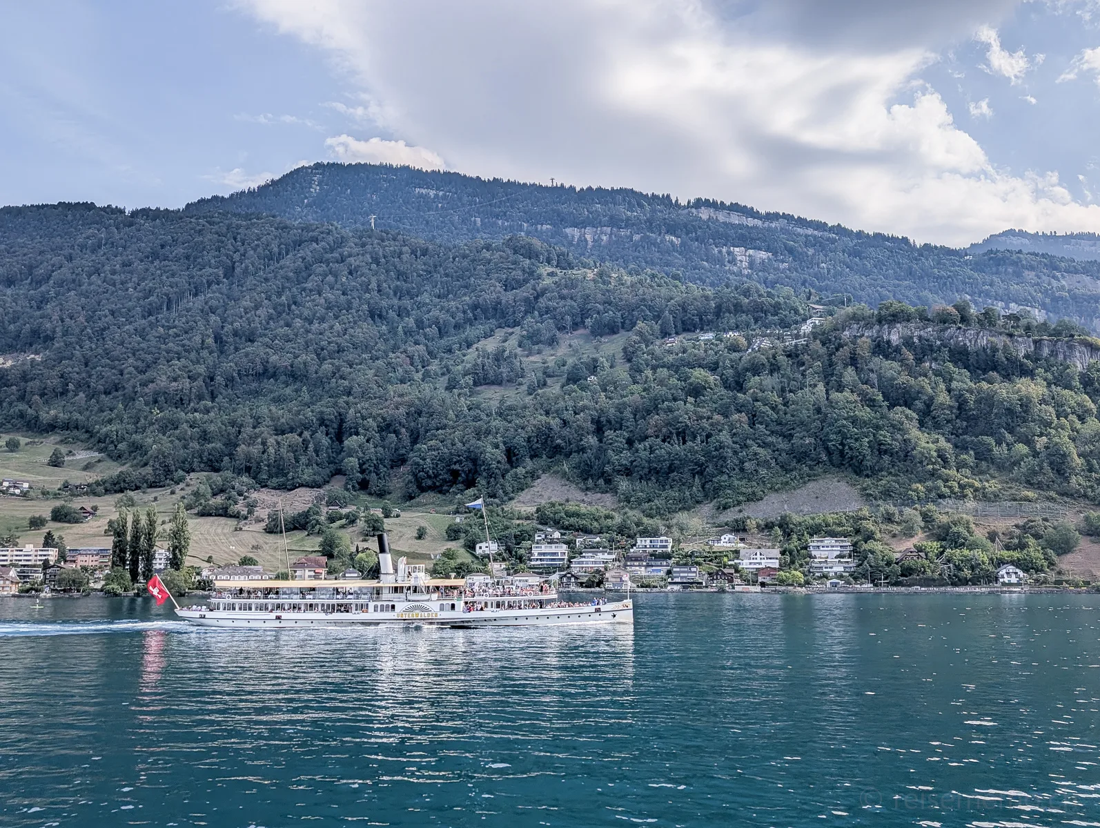 Ein Dampfschiff mit Schweizer Flagge fährt auf dem Vierwaldstättersee vor dem Ort Weggis, im Hintergrund grüne Berghänge