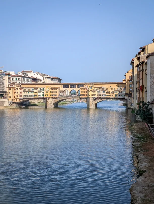 Blick auf den Ponte Vecchio über den Arno in Florenz mit seinen charakteristischen gelb-orangen Gebäuden auf der Brücke unter klarem blauem Himmel