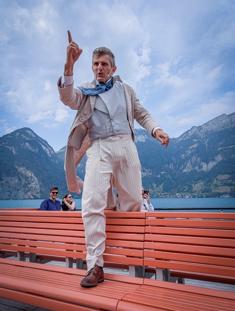 Ein Schauspieler in heller Kleidung performt gestikulierend auf orangen Bänken vor See und Bergen