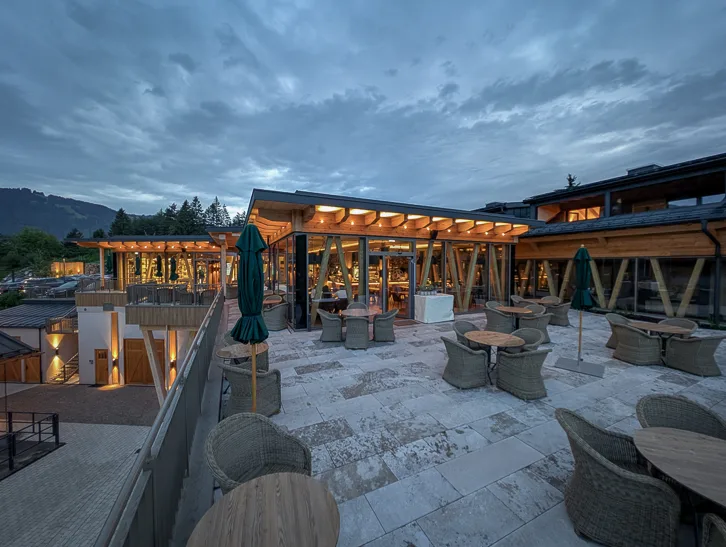 Weitläufige Aussentetrasse des Hofgut Restaurants im Sonnenalp Resort bei Abenddämmerung mit beleuchteten Holzstrukturen, Korbstühlen und Tischen vor alpiner Kulisse