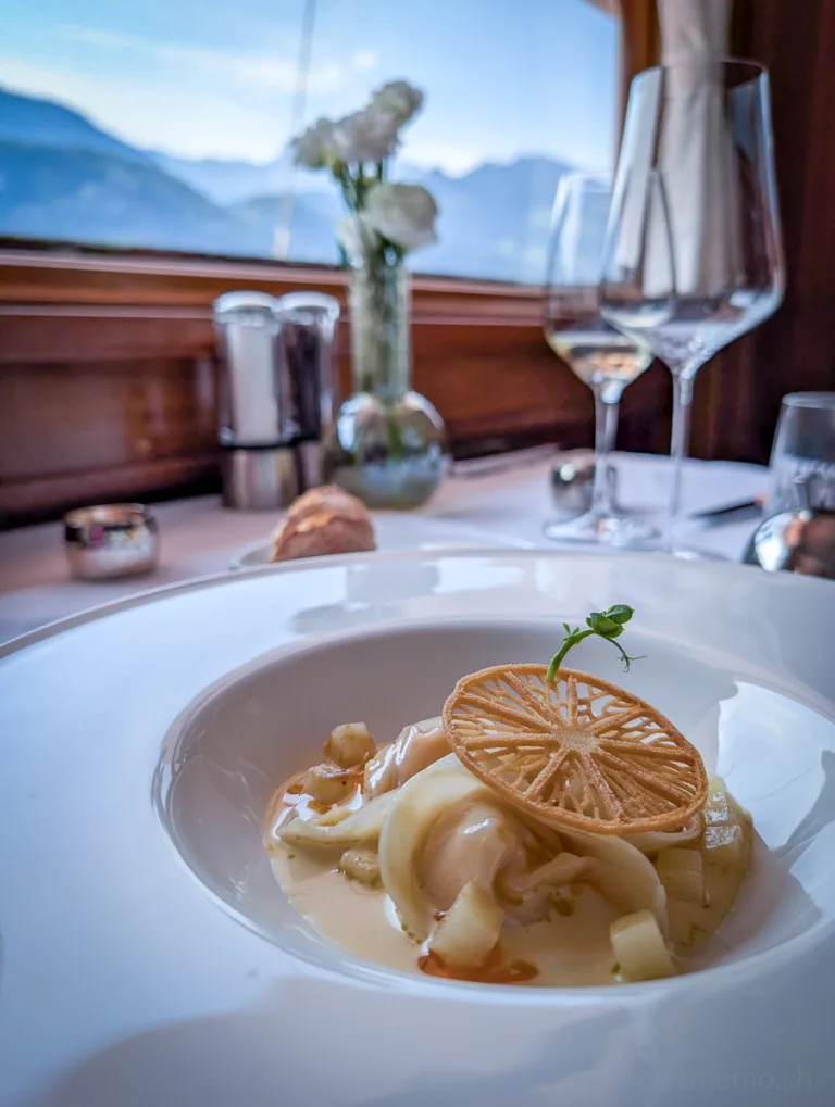 Weiss gedeckter Tisch mit Gourmet-Hauptgang in rundem Teller, dahinter Panoramablick auf Berge am Vierwaldstättersee