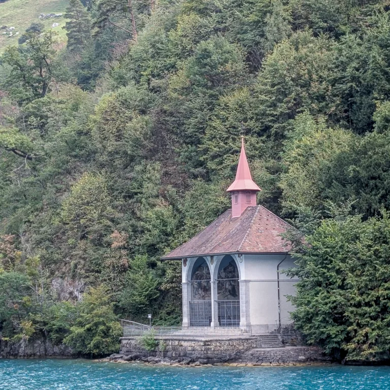 Eine kleine Kapelle mit spitzem Turm steht am Ufer des türkisblauen Vierwaldstättersees vor dichtem Wald