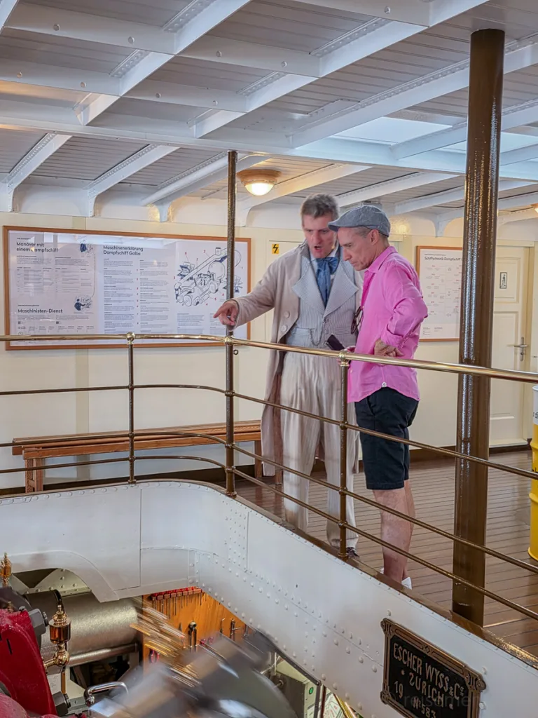 Zwei Männer stehen auf der Galerie eines historischen Dampfschiffs und blicken in den Maschinenraum