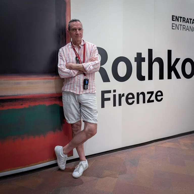 Walter Schärer lehnt mit verschränkten Armen an der Eingangsbeschilderung der Rothko-Ausstellung im Palazzo Strozzi in Florenz, neben einem grossformatigen abstrakten Gemälde in Rot, Schwarz und Grün