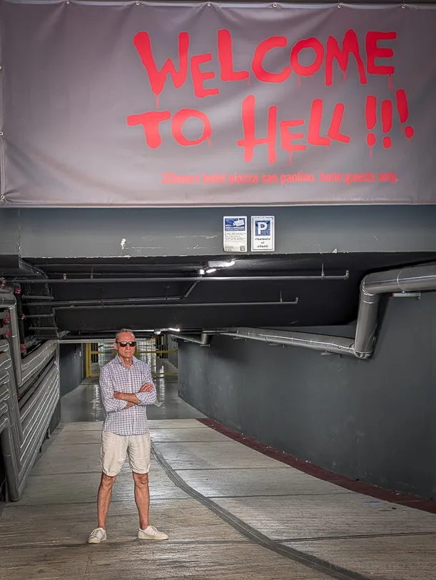 Ein Mann mit Sonnenbrille und verschränkten Armen steht vor der Einfahrt des 25hours Hotelparkhauses in Florenz, über ihm ein grosses graues Banner mit roter Aufschrift «Welcome to Hell»